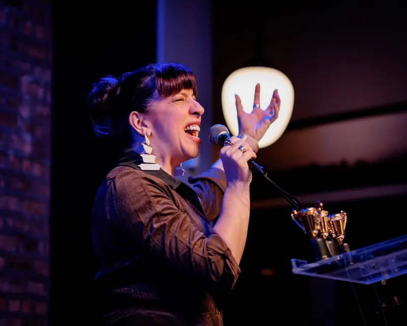 A performer laughing into the microphone, trophies on the lectern