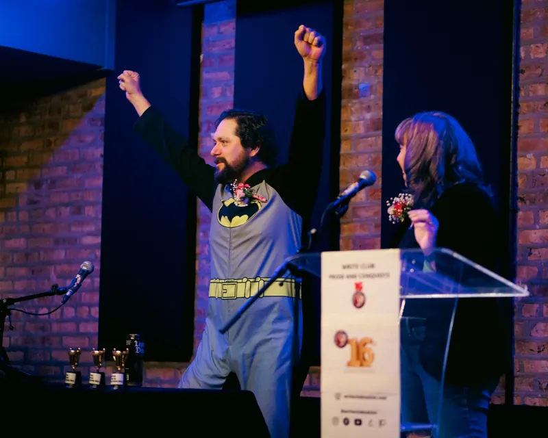 A combatant in a Batman onesie with both fists raised, the Prose and Conquests sign on the lectern
