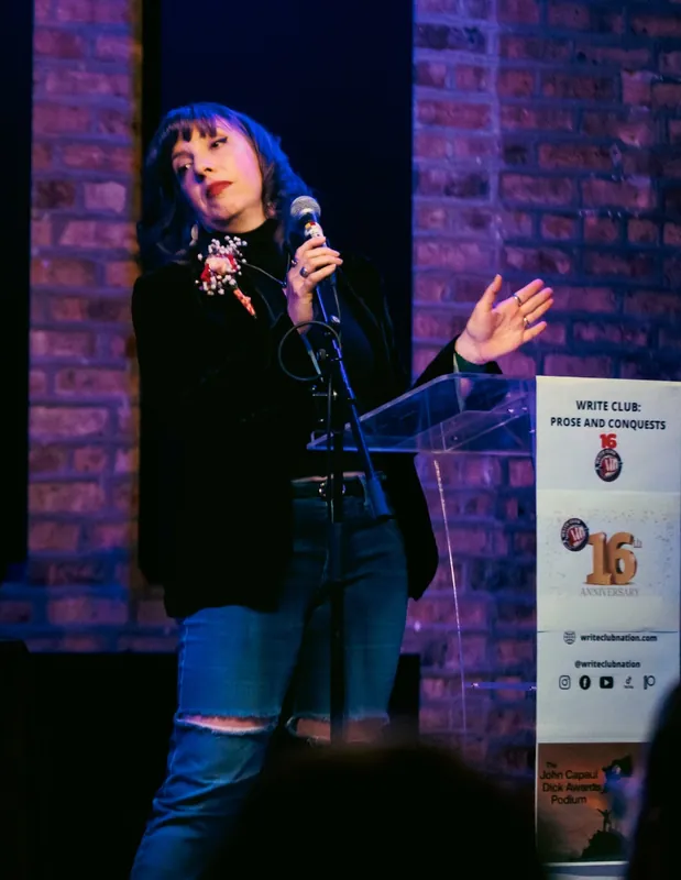 A combatant with a corsage, one hand raised at the microphone beside the Prose and Conquests sign
