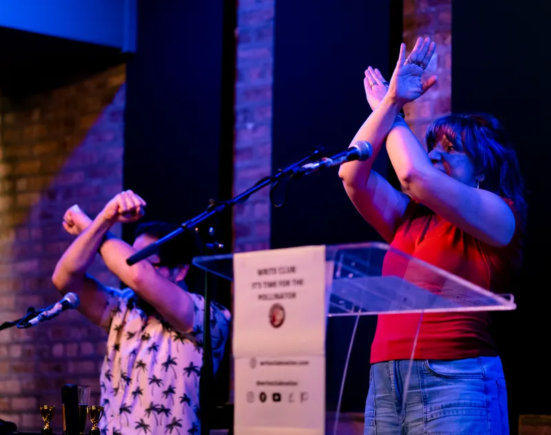 Two performers crossing their arms in an X at the lectern, the It's Time for the Pollinator sign below