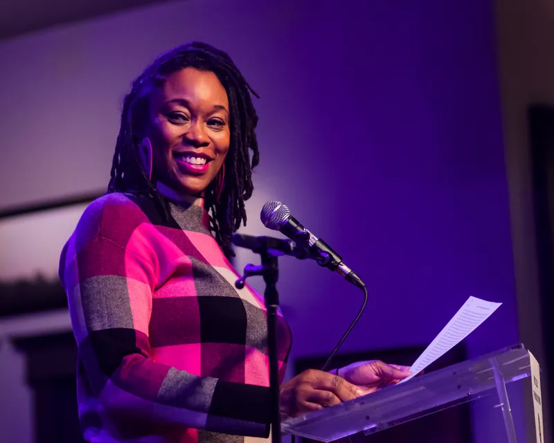 A combatant in a plaid top smiling from the lectern, pages in hand, against a purple wall