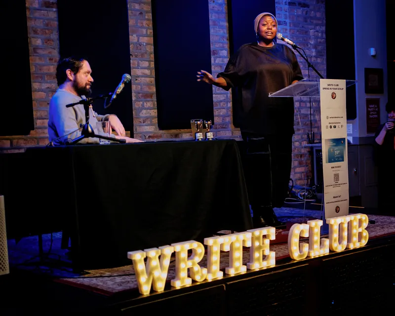 Wide shot of the stage, the timekeeper at the table, a combatant at the lectern and the lit WRITE CLUB marquee letters in front