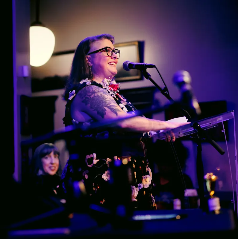 A combatant in glasses and a floral dress laughing at the lectern under a pendant lamp